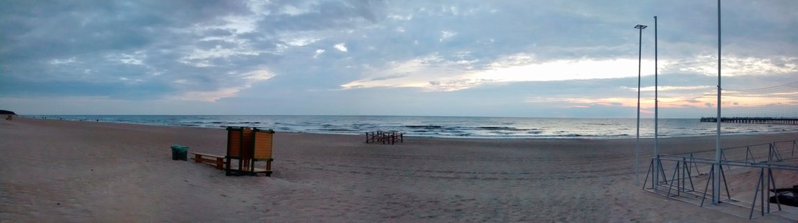 Wide beach scene showing sandy shore with wooden lifeguard tower, volleyball net poles, and pier in distance under cloudy evening sky at Palanga, Lithuania