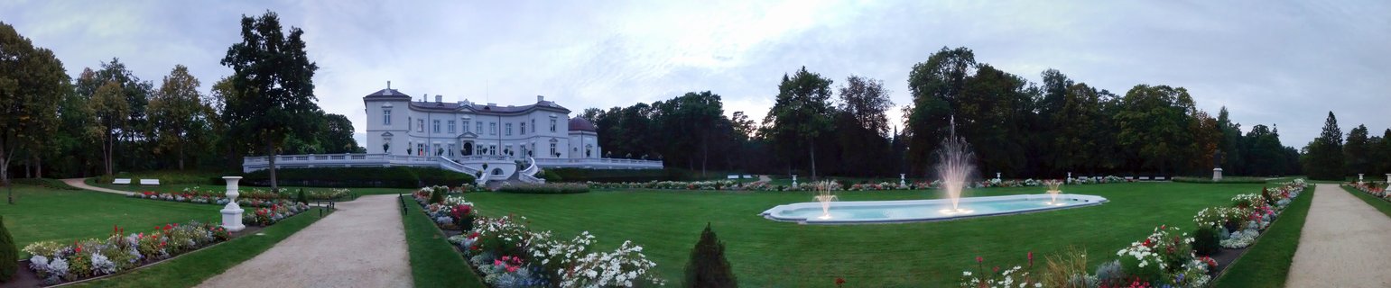 White neoclassical palace with formal gardens, flower beds, gravel pathways, and ornamental fountain under overcast evening sky in Palanga, Lithuania