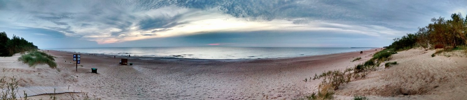 Panoramic beach scene showing wide sandy shore meeting calm Baltic Sea under cloudy sky, with sand dunes and vegetation on both sides