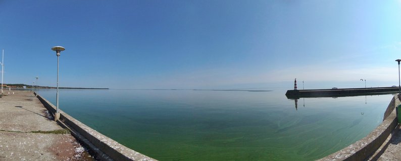 Panoramic view of a harbor with calm green-blue water, concrete pier walkway with street lamps on the left, and a red-and-white striped lighthouse on a breakwater extending from the right under a clear blue sky