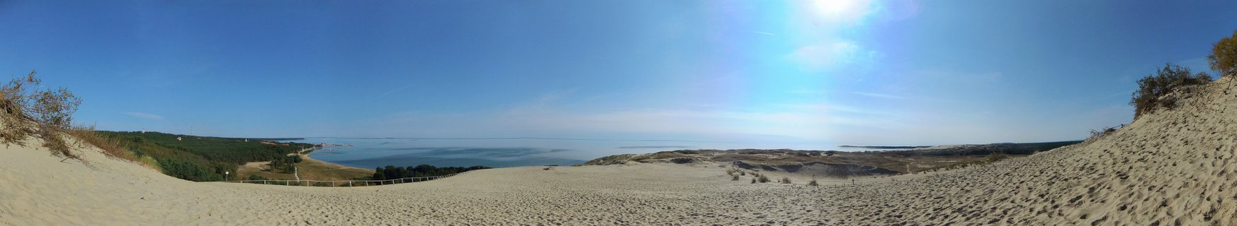 Wide panoramic view of sand dunes with Baltic Sea coastline visible in the distance under clear blue sky with bright sun