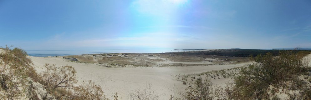 Wide coastal landscape showing white sand dunes with sparse vegetation, blue sky, and Baltic Sea horizon in Lithuania