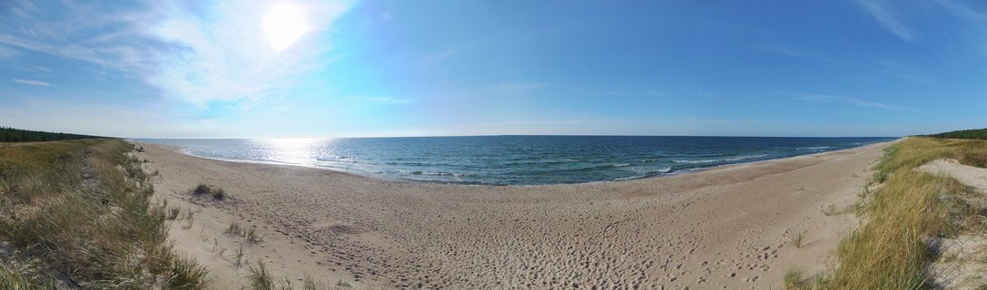 Wide coastal landscape showing a sandy beach with the Baltic Sea, grass-covered sand dunes on both sides, blue sky with wispy clouds, and sun reflecting on the water surface