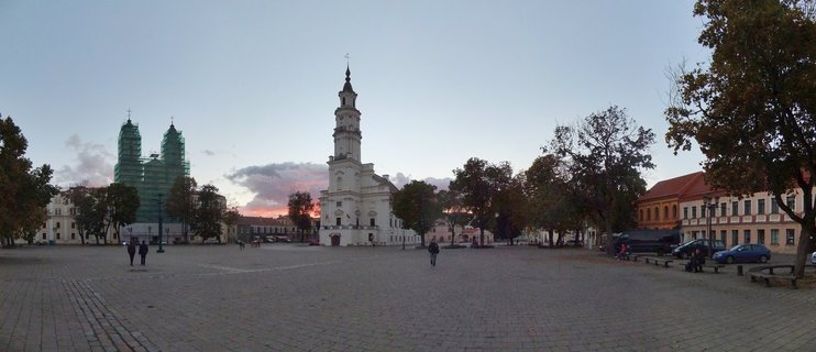 Panoramic view of Kaunas Town Hall Square at dusk showing white baroque church tower, cathedral with scaffolding, cobblestone plaza, and autumn trees under twilight sky
