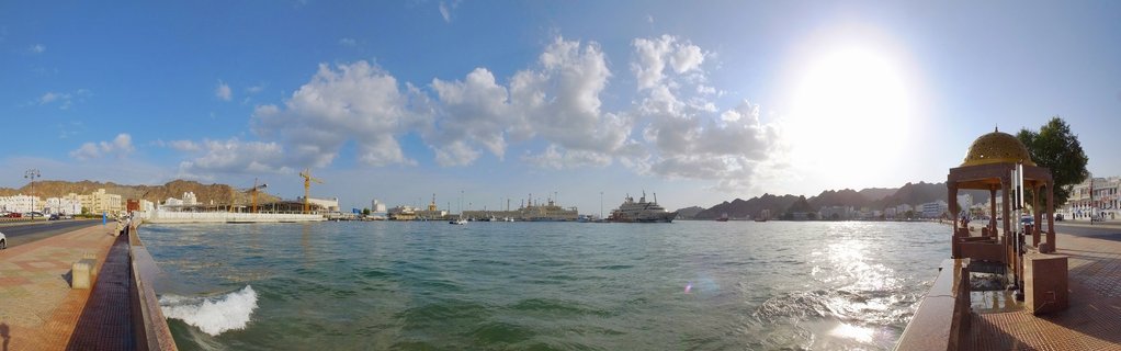 Waterfront harbor scene showing a golden-domed Islamic structure on a promenade, with ships docked in turquoise water, construction cranes, white buildings, and brown mountains under a bright sun with clouds in blue sky