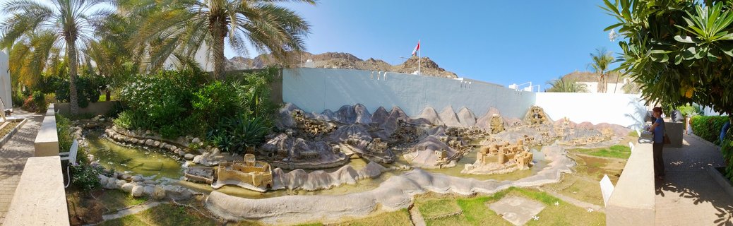 Outdoor miniature model park showing scaled replicas of Omani forts and landscapes with palm trees, water features, and walking paths under blue sky