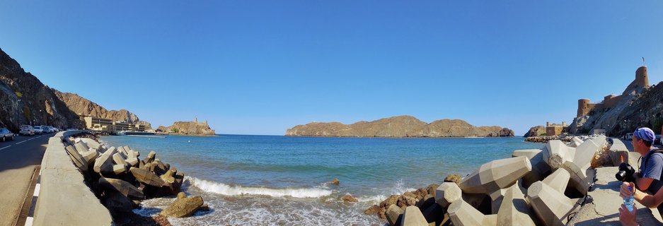 Panoramic coastal view of Muscat, Oman showing Al Jalali Fort on rocky outcrops, turquoise sea water with small waves, concrete breakwater blocks, coastal road with vehicles, and a photographer on the right side under clear blue sky