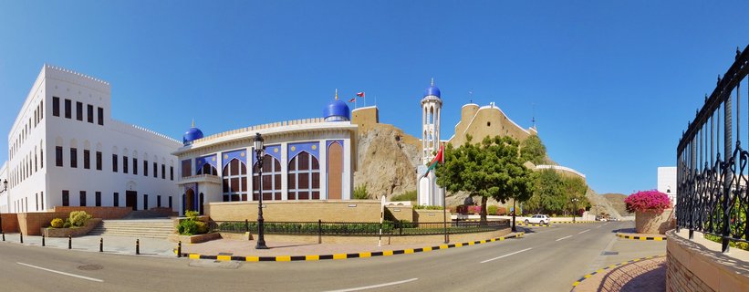 White government building with blue Islamic domes and minaret tower with historic stone fort on rocky hill in background under clear blue sky in Muscat Oman