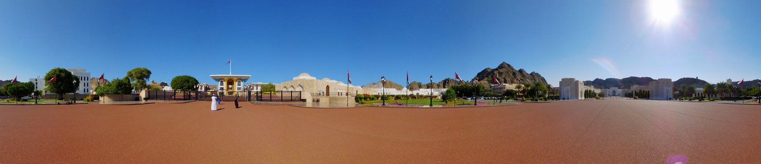 Wide view of Al Alam Palace in Muscat Oman with cream-colored traditional buildings, Omani flags on poles, two people in white traditional dress, rocky mountains in background, clear blue sky, large open plaza with reddish ground
