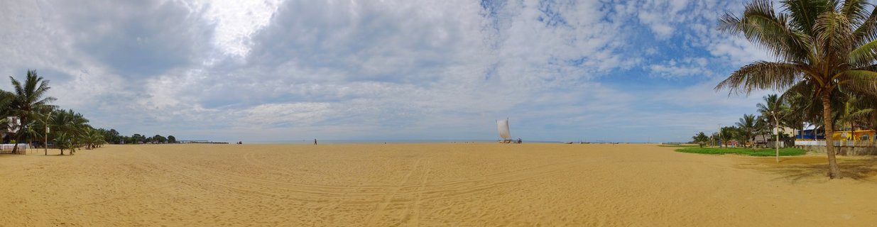 Wide panoramic beach scene showing golden sand, palm trees on both sides, a fishing boat with white sail on the water, and blue sky with clouds