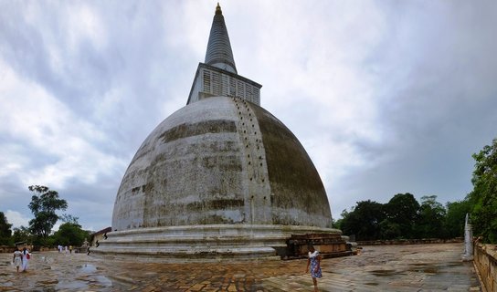Large white Buddhist stupa with conical spire standing on wet stone courtyard with people walking nearby under cloudy sky in Anuradhapura Sri Lanka