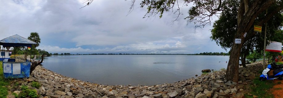 Wide waterfront view showing a large reservoir with rocky stone embankment in foreground, blue kiosk structure on left, large tree on right, and cloudy sky above calm water