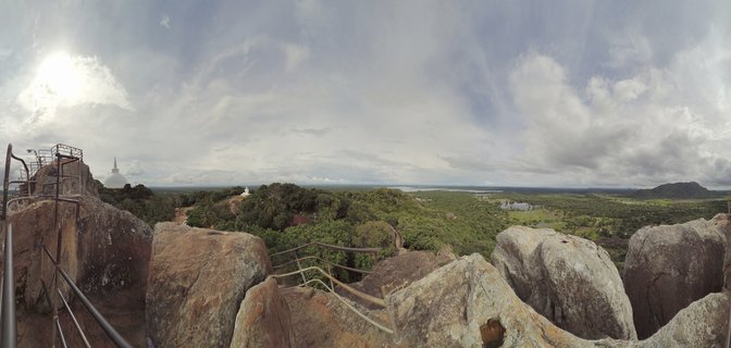 Wide panoramic landscape showing rocky foreground with metal railings, ancient white stupa rising above green trees, and distant water body under cloudy sky in Polonnaruwa, Sri Lanka