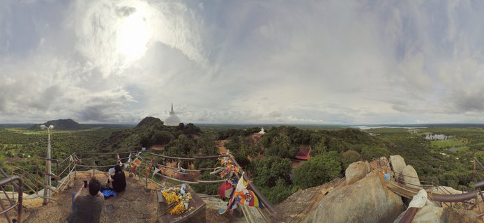 Panoramic landscape view from rocky summit showing white Buddhist stupa in distance, tropical green forest, prayer flags, metal railings, and two people sitting on rocks under cloudy sky in Sri Lanka