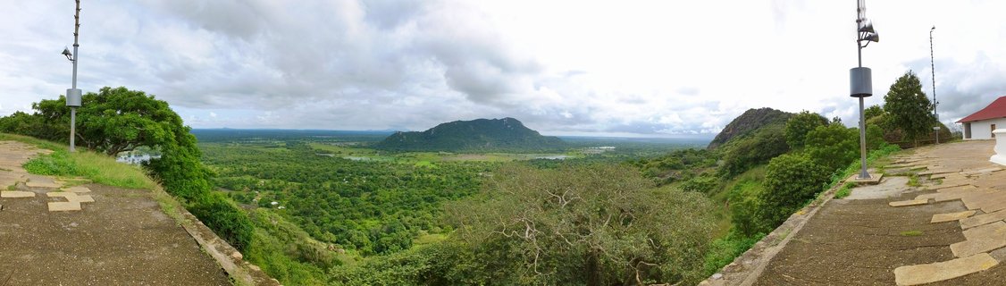 Wide panoramic landscape showing green forested plains and hills viewed from an elevated concrete platform with metal poles and a white building visible at the edges