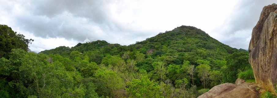 Panoramic landscape showing a white Buddhist stupa dome on a green forested hill with large rock formations in the foreground under cloudy sky in Polonnaruwa, Sri Lanka