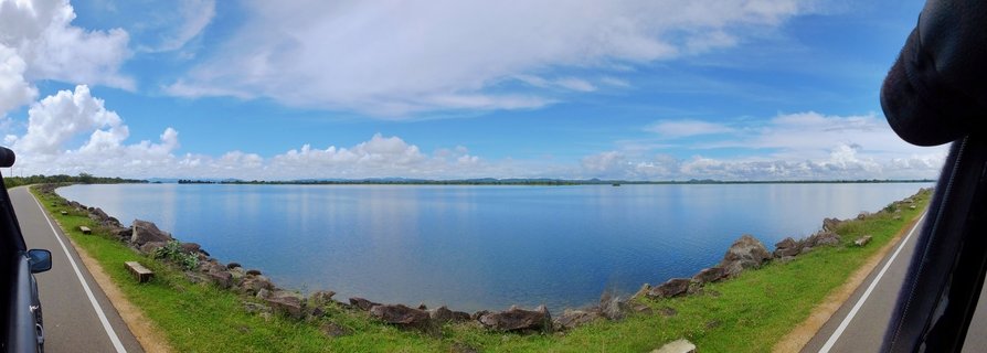 Wide panoramic view of a large reservoir with calm blue water, rocky embankment with green grass, paved road, and blue sky with white clouds in Polonnaruwa, Sri Lanka
