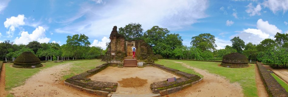 Ancient stone temple gateway ruins with steps leading to an entrance where a person stands, flanked by two dome-shaped stupas on a grassy courtyard surrounded by trees under blue sky with clouds.