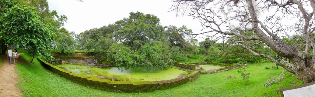 Ancient brick ruins with moss-covered water pool surrounded by lush tropical vegetation and large trees at Polonnaruwa UNESCO World Heritage Site in Sri Lanka, with two tourists walking on a dirt path under overcast sky