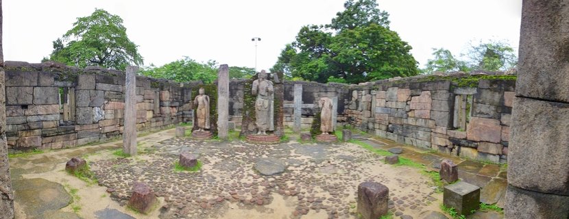 Three standing Buddha statues positioned within ancient stone wall ruins at an archaeological site with cobblestone ground and trees visible above the walls
