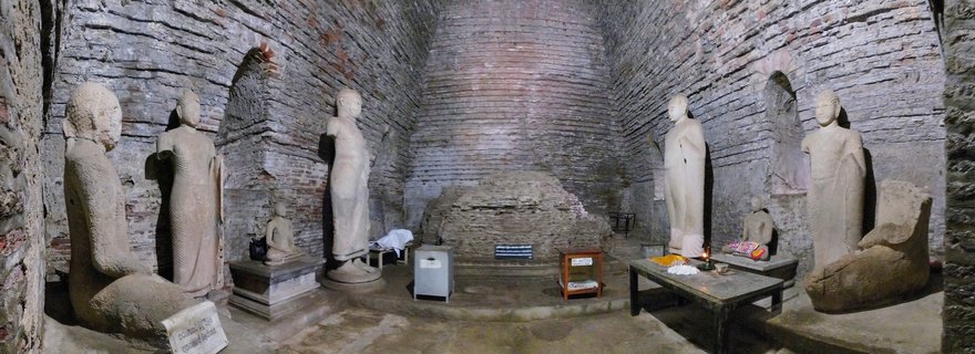 Interior of ancient Buddhist temple showing multiple stone Buddha statues in standing and seated positions along weathered brick walls with a central stupa structure