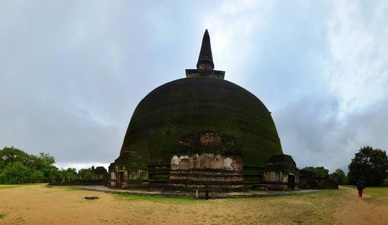 Large moss-covered Buddhist stupa with conical spire in ancient archaeological site with sandy ground and overcast sky