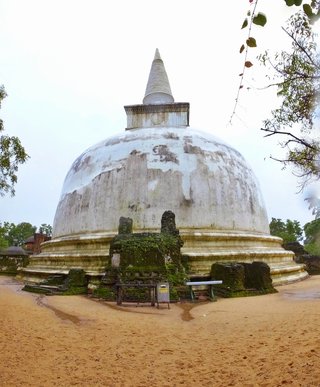 Large white dome-shaped Buddhist stupa with weathered plaster and conical spire, surrounded by moss-covered ancient brick ruins on sandy ground with trees framing the sides under overcast sky