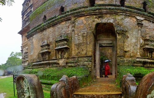 Ancient circular brick stupa with moss-covered walls, arched doorway, and carved stone guardstones in foreground. Buddhist monk in red robes holding umbrella enters doorway during rain. Overcast sky, wet stone steps, tropical vegetation visible.