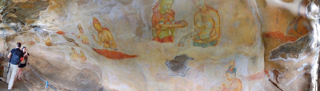 Ancient Buddhist frescoes painted on a rock cave wall showing multiple figures in traditional attire with jewelry, with two tourists viewing the artwork