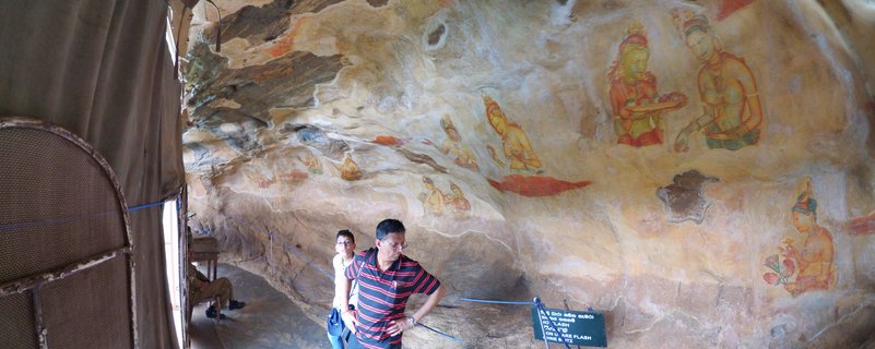 Ancient rock wall frescoes showing painted female figures in traditional dress at Sigiriya archaeological site with visitors viewing the artwork