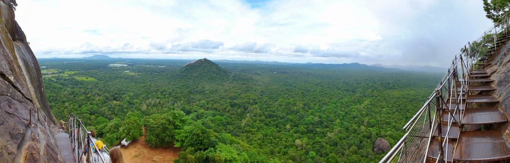 Wide panoramic photograph showing jungle landscape viewed from elevated rock position with metal staircases and railings visible on both sides, dense green forest canopy extending to horizon with rock formation in distance under cloudy sky