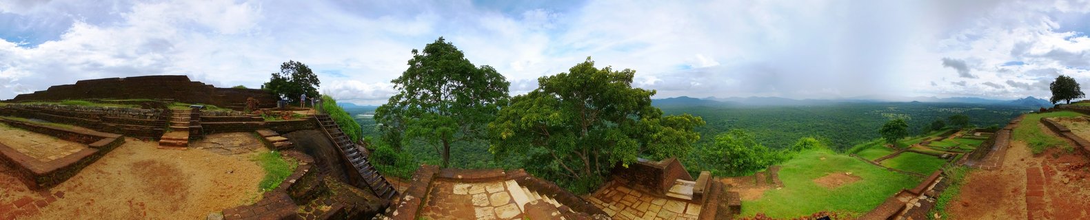 Panoramic photograph showing ancient brick ruins and stone terraces on top of Sigiriya rock with green tropical forest and distant mountains visible in the background under a partly cloudy sky