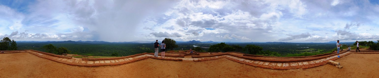 Panoramic view from Sigiriya Rock Fortress summit showing tourists on ancient brick terrace overlooking green landscape with mountains and cloudy sky