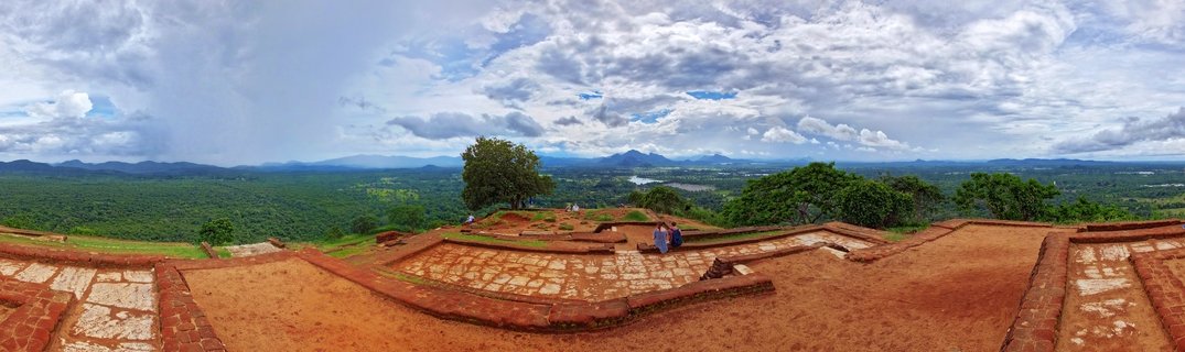 Panoramic view from the top of Sigiriya Rock Fortress showing ancient brick terraces with tourists, overlooking green jungle landscape and distant mountains under cloudy sky in Sri Lanka