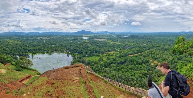 Panoramic landscape view from elevated rock formation showing two tourists at railing overlooking vast green tropical forest with lakes and distant mountains under cloudy sky in Sigiriya, Sri Lanka