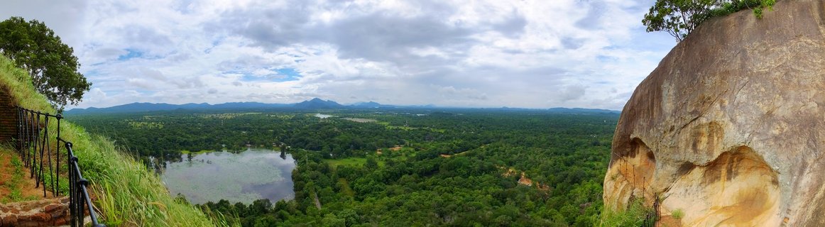 Panoramic landscape view from elevated rock formation showing forest, lake, mountains, and cloudy sky with metal safety railing visible on left side