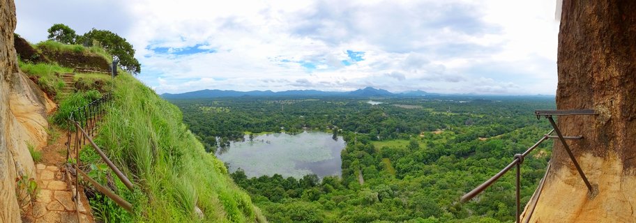 Panoramic landscape view from Sigiriya Rock Fortress showing green tropical forests, a lotus-covered lake, stone steps with metal railings, and distant mountains under cloudy sky in Sri Lanka