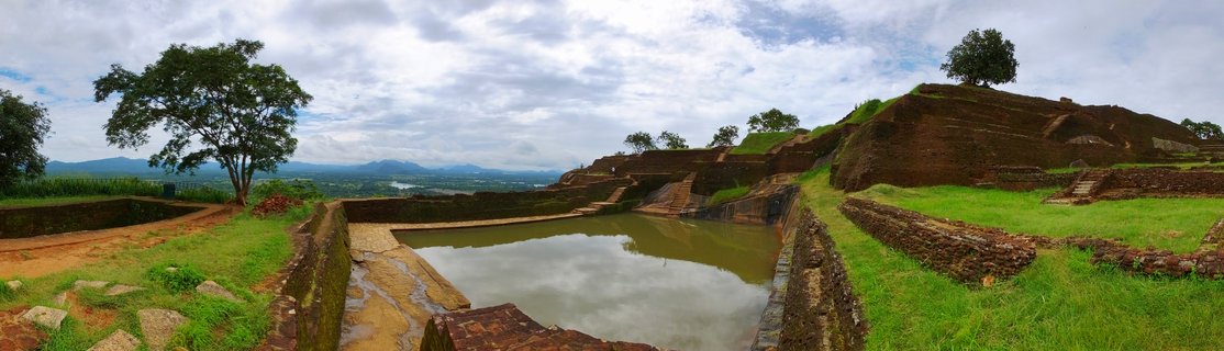 Ancient stone water cistern surrounded by brick terraces and green grass at Sigiriya rock fortress archaeological site in Sri Lanka with distant mountains under cloudy sky