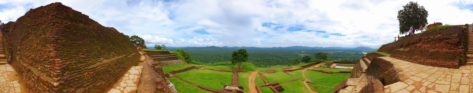 Panoramic photograph showing ancient brick ruins and stone terraces on top of Sigiriya rock with green jungle landscape and distant mountains under partly cloudy sky