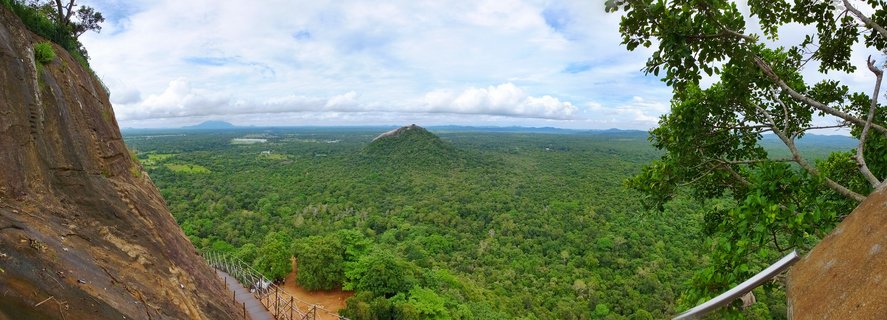 Panoramic view from Sigiriya Rock Fortress summit showing tropical jungle canopy stretching to horizon, with Pidurangala Rock visible in middle distance, granite cliff face and metal safety railing in foreground, partly cloudy sky above, Central Province, Sri Lanka