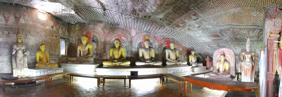 Interior view of Dambulla Cave Temple showing multiple golden and white Buddha statues in seated meditation positions arranged along stone platforms, with ancient painted ceiling frescoes featuring geometric patterns in red, white and black colors covering the natural rock cave ceiling