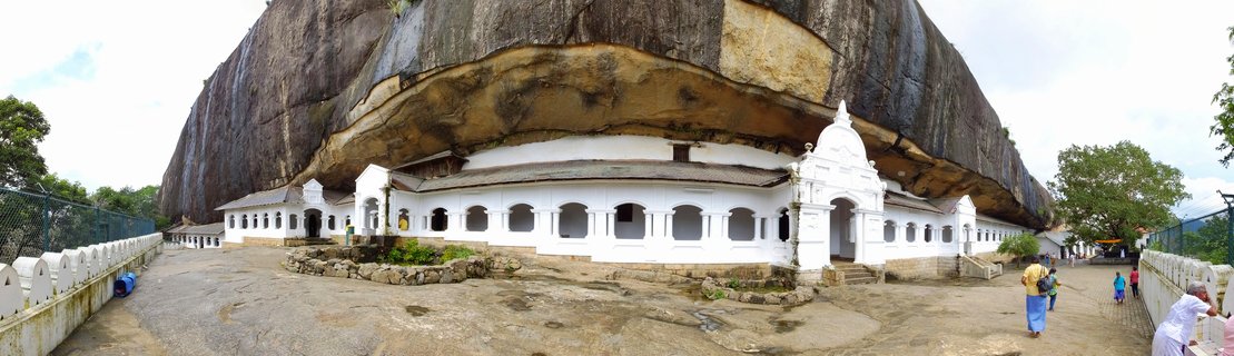 White Buddhist temple buildings with arched windows built beneath a massive curved rock overhang at Dambulla Cave Temple in Sri Lanka, with visitors walking on stone courtyard