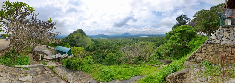 Panoramic landscape view from Dambulla showing stone walls and pathways in foreground, lush green tropical valley, distinctive conical hill, and distant Sigiriya rock under cloudy sky
