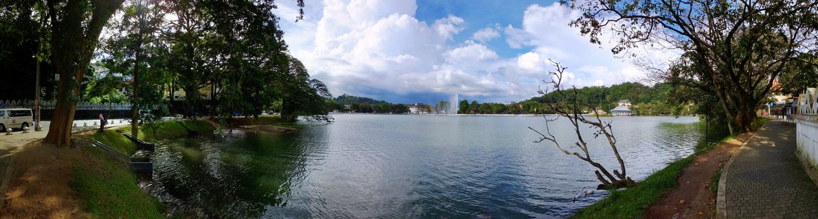 Wide panoramic photograph of Kandy Lake showing a central fountain spraying water, with tree-lined walkways on both sides and buildings visible in the background under a blue sky with white clouds.