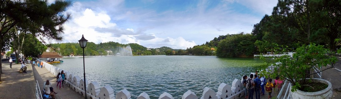 Wide view of Kandy Lake in Sri Lanka showing a central fountain, white decorative wall barrier, visitors on promenade, surrounding trees and hills under blue sky with clouds
