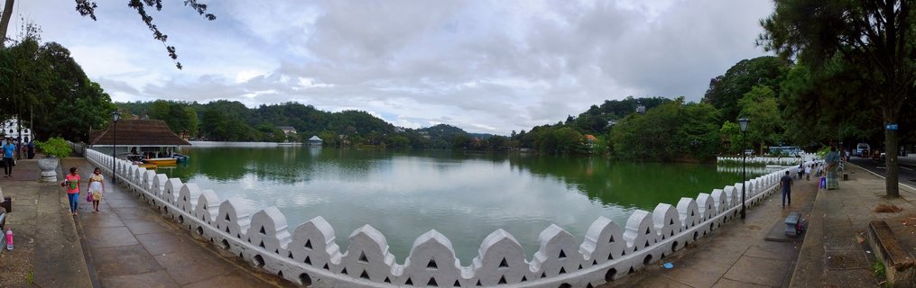 Panoramic view of Kandy Lake surrounded by white decorative wall with triangular patterns, people walking on paved pathway, green water, trees and hills in background under cloudy sky