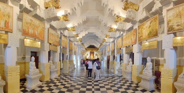 Interior corridor of a Buddhist temple with black and white checkered floor, white pillars with golden decorations, Buddha statues on pedestals, religious murals on walls, and visitors walking through the hall