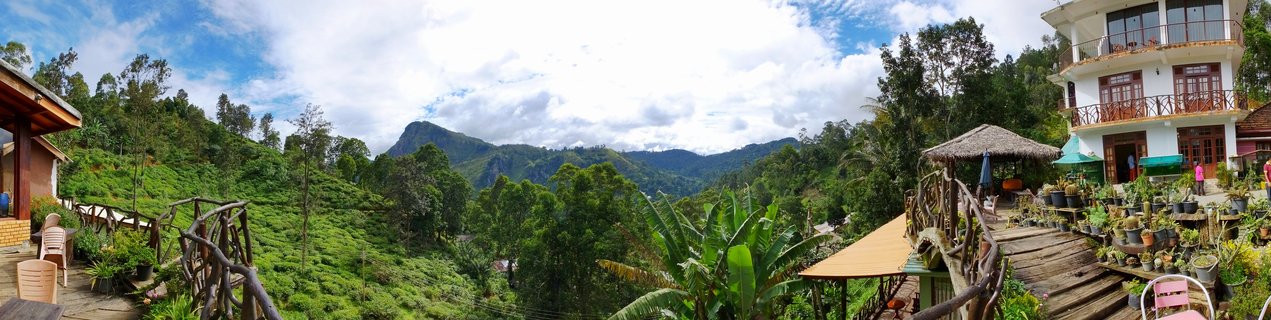 Panoramic view from a guesthouse terrace showing green mountains, tea plantations, white buildings with wooden balconies, and tropical vegetation in Ella, Sri Lanka
