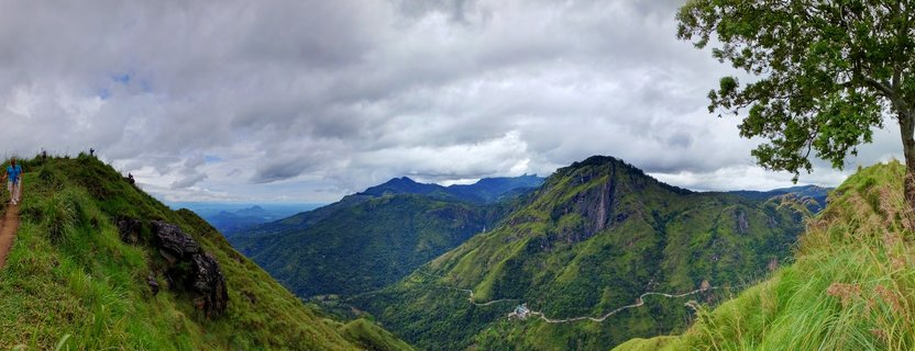 Panoramic landscape showing green mountain ranges with hiking trail on the left, winding road in valley below, and cloudy sky over Ella, Sri Lanka