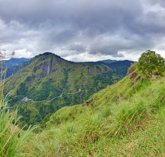 Panoramic view of green mountains in Ella, Sri Lanka with a winding road through the valley under cloudy sky